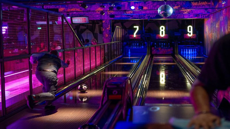 Enjoy thrilling arcade games, bowling, darts, crazy golf and more at Lane7 London Victoria. Image courtesy of Lane7 London Victoria. activity bar focusing on a bowling alley with someone throwing the ball, with pink and purple graffiti on the walls and a disco ball glistening