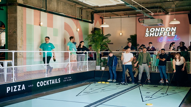 Indoor and outdoor shuffleboard courts at London Shuffle Club. Image courtesy of London Shuffle Club. An open planned room with wooden panelling and shuffleboard courts, with two teams playing a game and having fun.