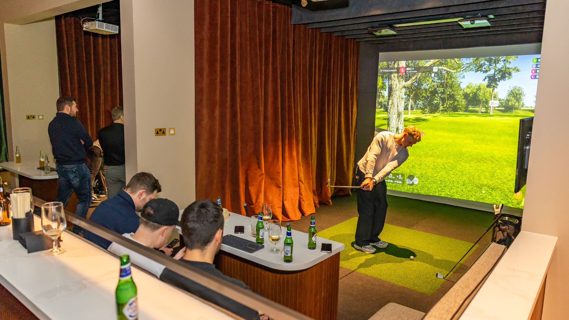 Experience world-class indoor golf with Trackman-powered bays at Pitch Canary Wharf. Image courtesy of Pitch Canary Wharf. a large golf simulator bay with a man swinging a golf club, with three men sat on a plush sofa holding a beer and watching