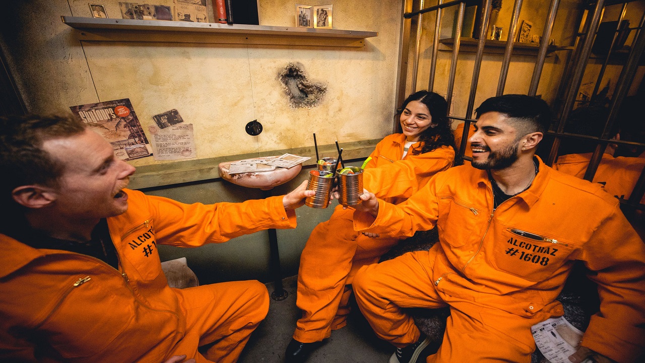 Enjoy an immersive experience at the Alcotraz Immersive Prison Cocktail Experience in London. Image courtesy of Golden Tours. three people wearing orange prison jumpsuits, sat down in a cell, smiling and holding their cocktails up in the air