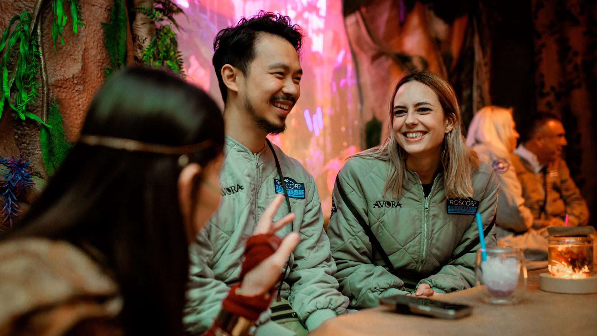 Explore the magical world of Avora and interact with the local inhabitants in Avora: New World Cocktail Experience. Image courtesy of Golden Tours. two people wearing jumpsuits and sat at a table, smiling and interacting with actors in the immersive experience