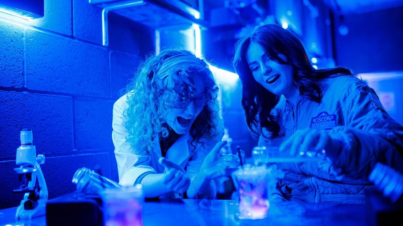 Start your Avora journey in the Roscorp Laboratory and create a colour-changing antidote cocktail. Image courtesy of Golden Tours. two women in jumpsuits, one an actor playing a doctor who is helping the other woman to create a magical cocktail
