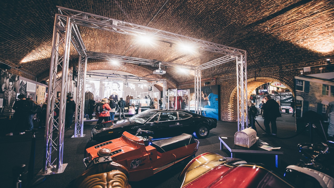 See famous memorabilia at the Direct from Graceland: Elvis exhibition, including the famous Ferrari Dino GT4. Image courtesy of Golden Tours. large exhibition space with exposed brickwork ceilings and walls, and people walking around the displays of cars and Elvis Presley memorabilia