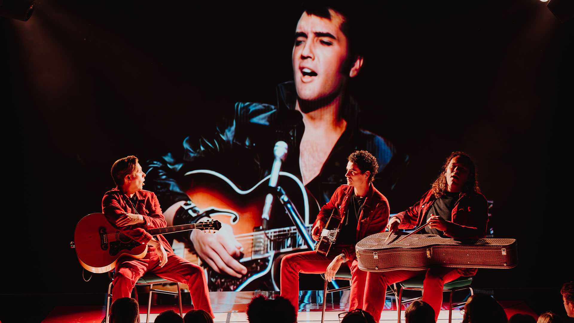 Immerse yourself in Elvis Presley's 1968 Comeback Special performance at the new immersive experience, Elvis Evolution, in London. Image courtesy of Layered Reality. At Elvis Evolution, a projection of Elvis Presley is shown on a screen, accompanied by a three-piece band playing various instruments.