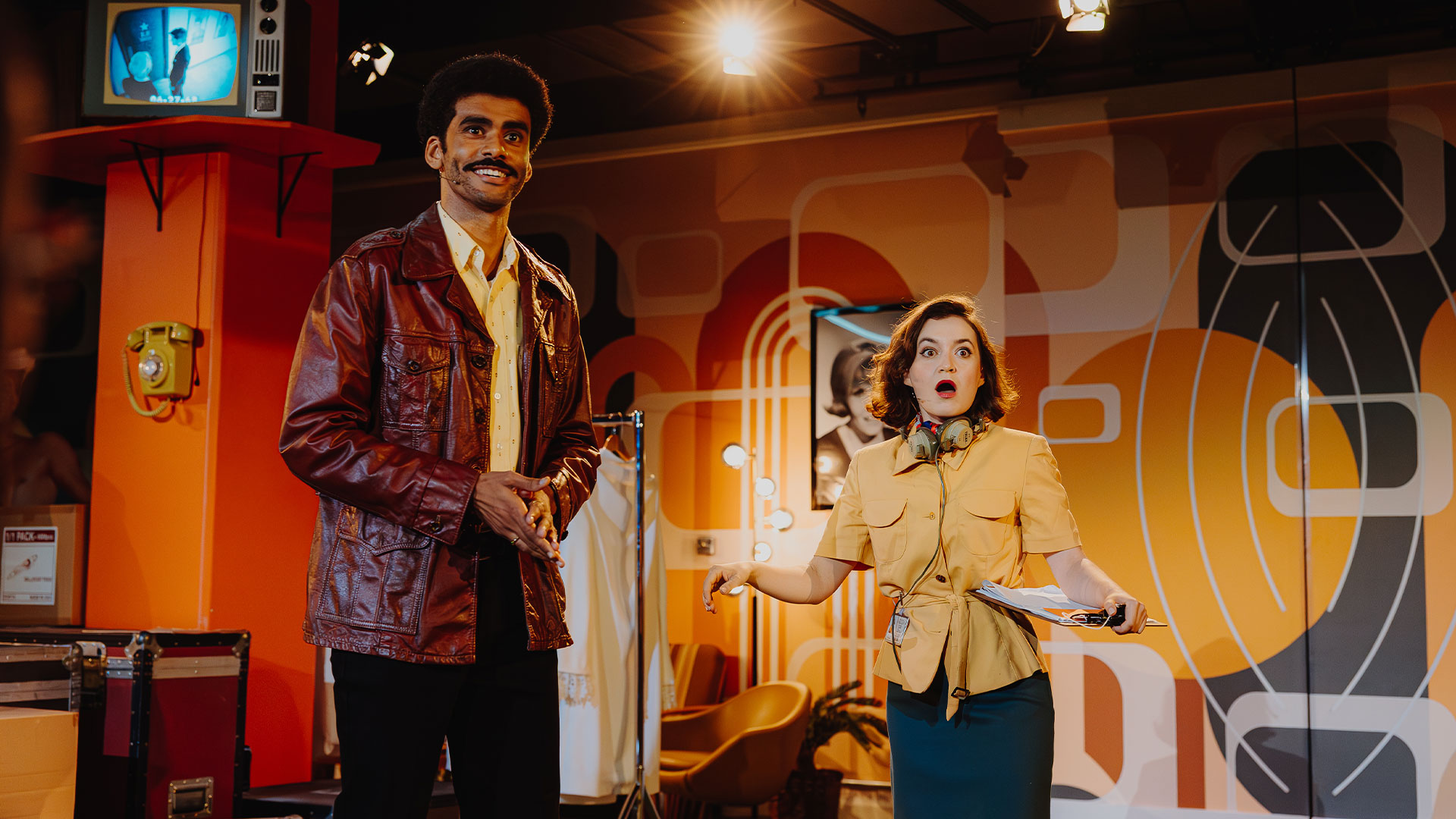 Visit the new London immersive experience, Elvis Evolution, and discover the '68 Comeback Special, hosted at NBC Studios. Image courtesy of Layered Reality. The studio operator is wearing a yellow jacket and denim skirt and looks shocked. Next to her is the actor playing Elvis's best friend, who is wearing a brown leather jacket and has a big smile on his face.