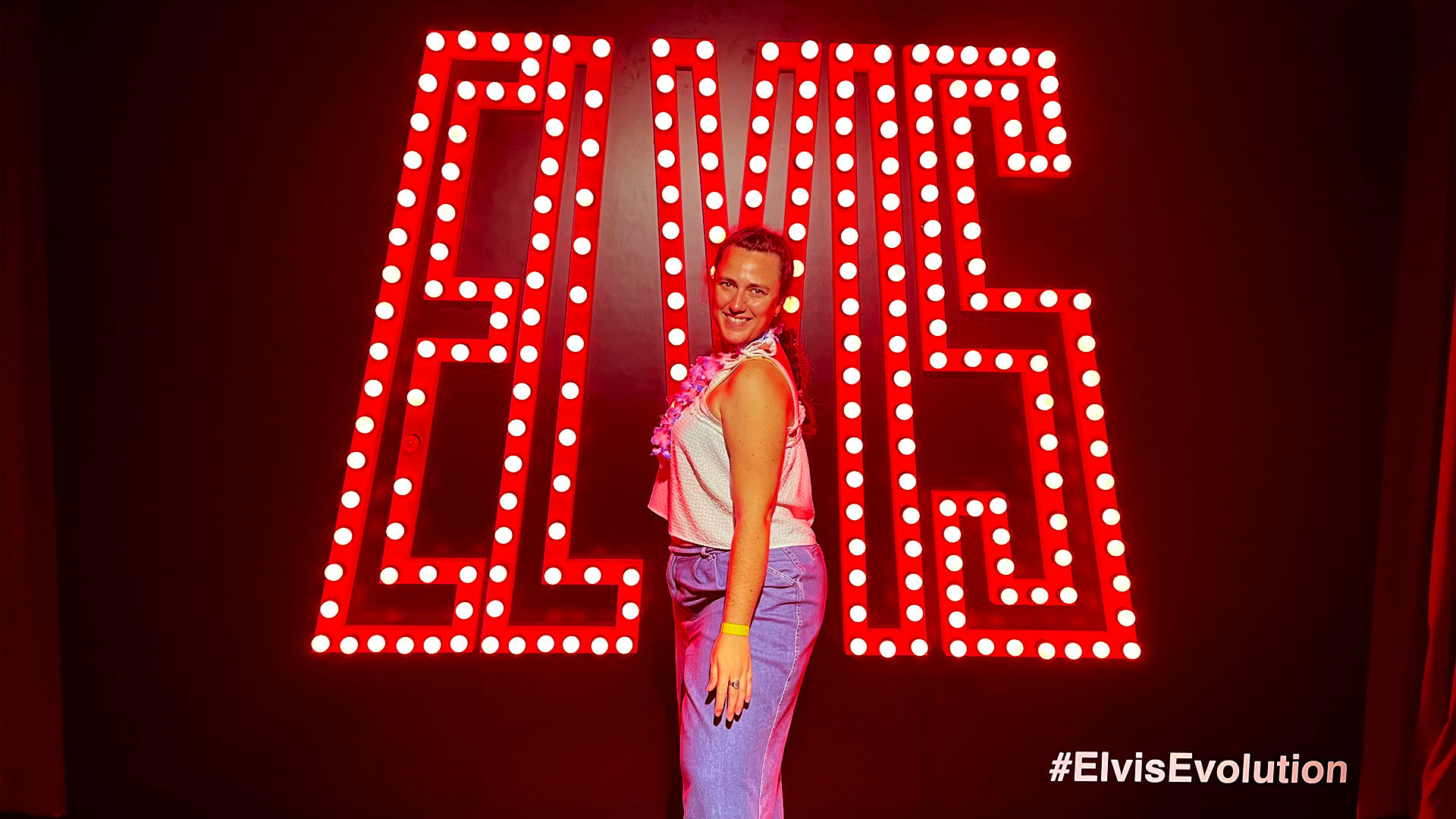 Make sure you don't miss any photo opportunities at the All Shook Up Party, which is part of the immersive Elvis Evolution experience. Image courtesy of Carole Rocton. A woman wearing a white top and jeans stands in front of a red neon sign spelling out the name 'Elvis'.