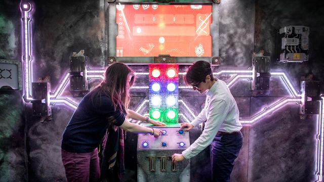 clueQuest Origenes escape room. Photo: David Levene. Image courtesy of clueQuest. Two people stand by a control board with multi-coloured lights and buttons in a london escape room.