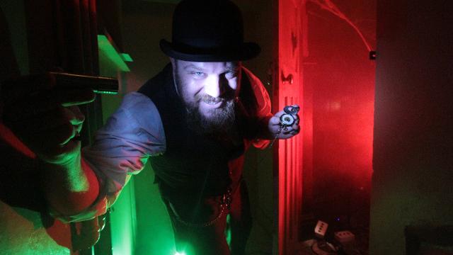 Can you uncover the sordid secrets of Lady Chastity? Image courtesy of Lady Chastity's Reserve A man in a black bowler hat stands under red and green light holding an escape room clue.