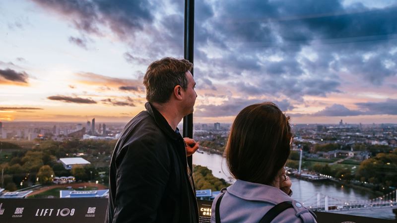 Experience Lift 109 at Battersea Power Station. Credit: Joshua Atkins. Image courtesy of Lift 109. a man and a woman looking out of a window with a view of London's skyline