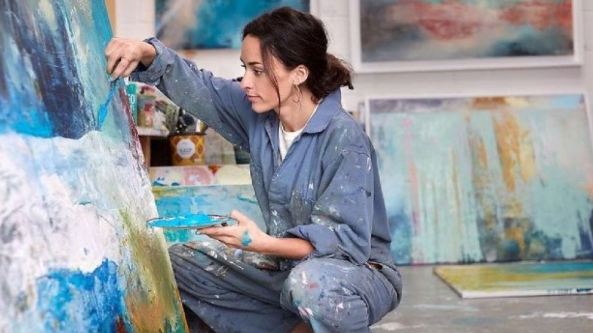 Painting class in London. Image courtesy of Red Letter Days. Woman painting on a canvas using blue, white and yellow paint, with paintings in the background.