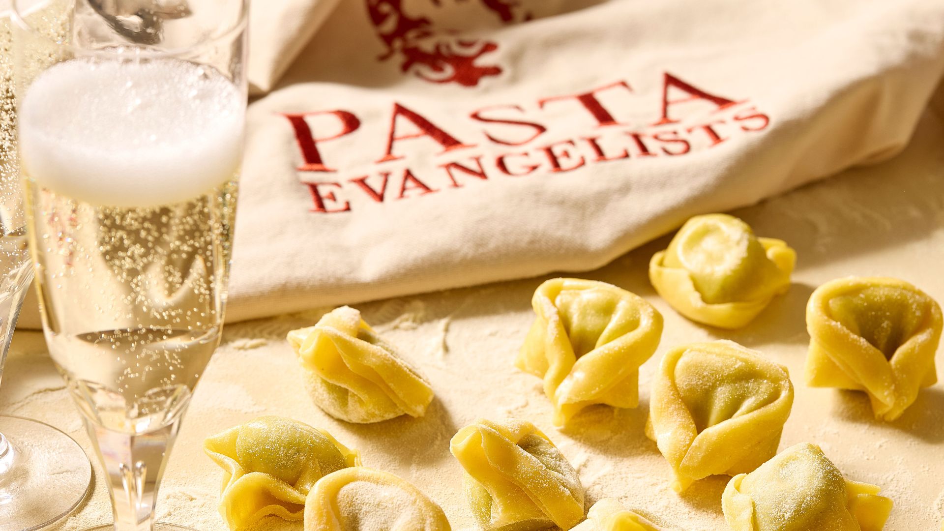 Join a pasta-making masterclass at Pasta Evangelists. Image courtesy of Red Letter Days. close up of small ravioli pasta on a flour workbench, with a Pasta Evangelists bag in the background and a glass of bubbly