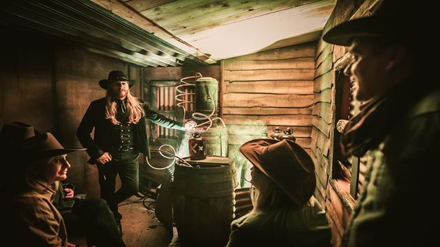 A dark shed-style room, with a distillery in the background, and people huddled around in the room looking at it