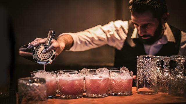 A bartender pouring cocktails into small tumble glasses lined up in a row