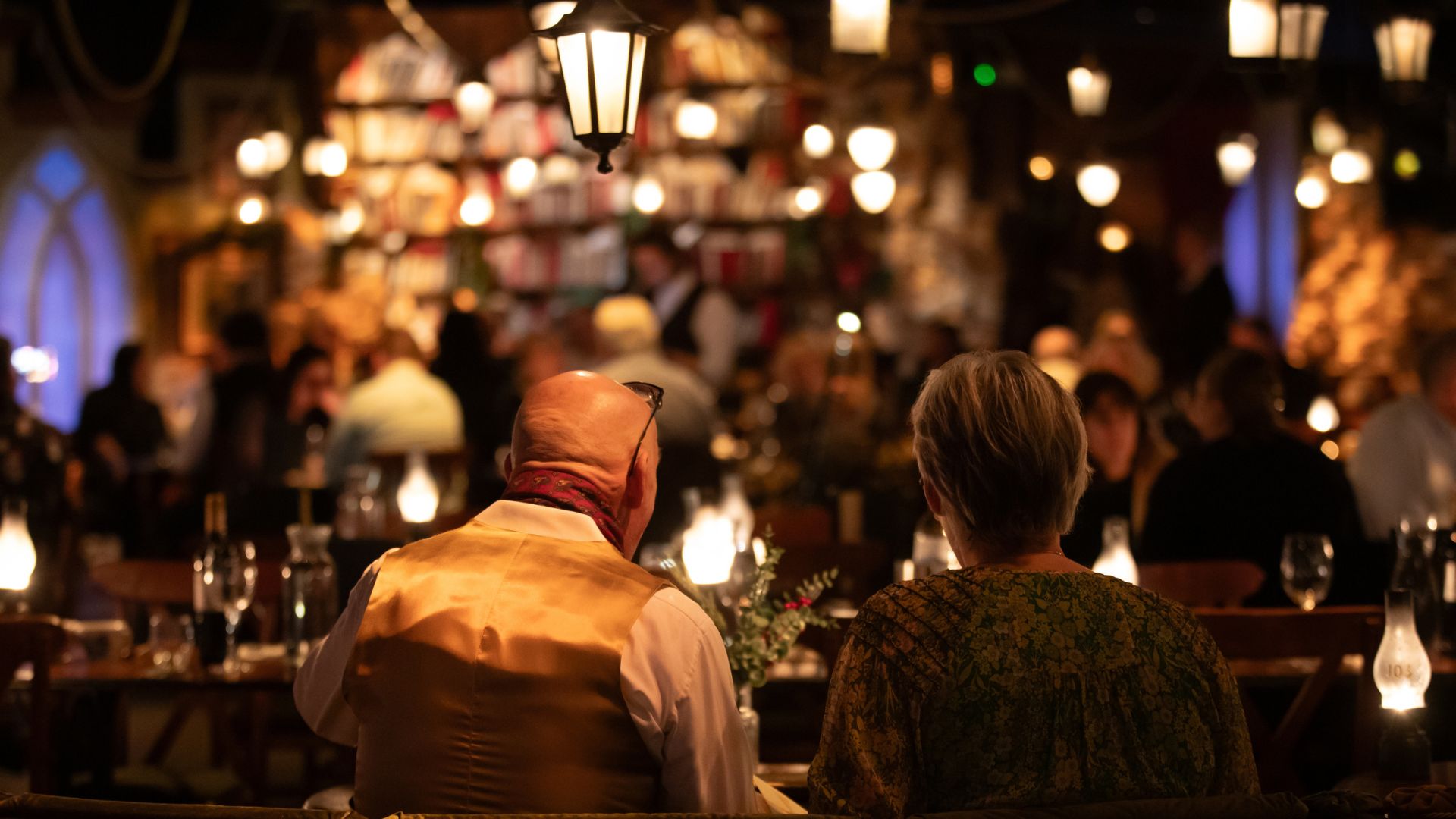 Settle into your seats at the festive immersive dining experience, The Great Christmas Feast. © Amy Williams Consultancy/Ali Wright two people sat at a table, look away from the camera in a dim fairy lit room