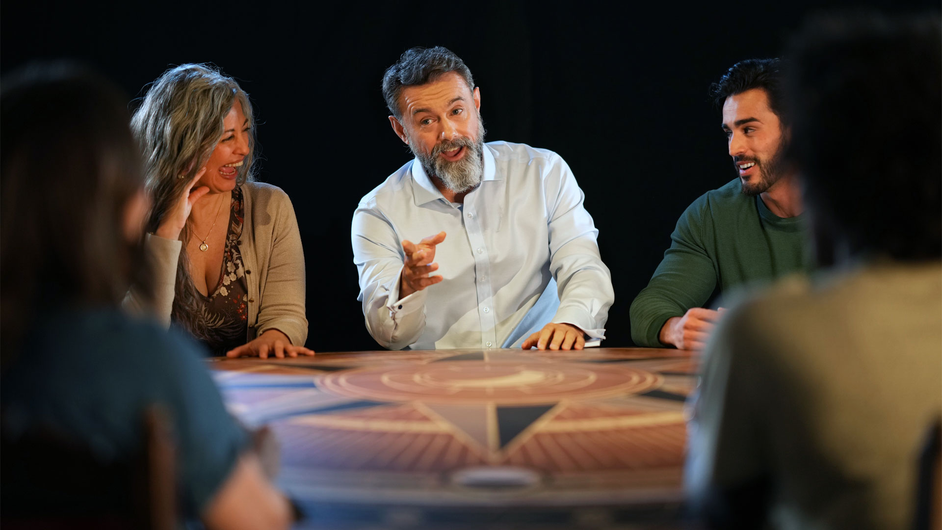 Play the official live game of the TV phenomenon at The Traitors: Live Experience. Image courtesy of Mance Communications. Photo credit: Hugo Glendinning. Group of people sitting around 'the Round Table' with man in centre pointing to person on the opposite end.