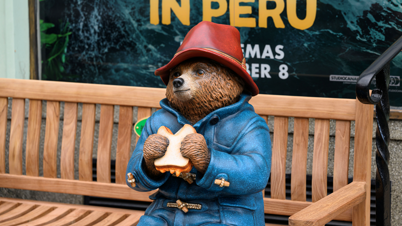 A statue of Paddington bear with his trademark blue coat and red hat sat on a wooden bench holding a marmalade-filled sandwich