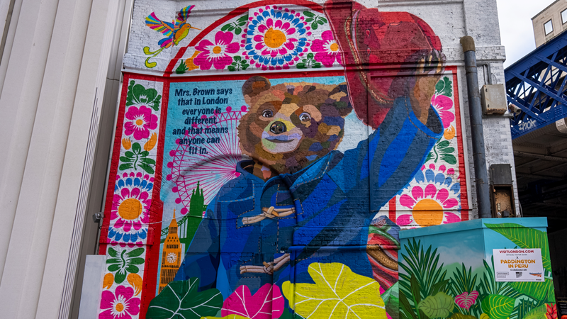 Head to Paddington's vibrant London mural on Sutton Walk outside Waterloo station. Credit: Peter Cohen/London & Partners. Image courtesy of London & Partners. A photo of a colourful painted mural of Paddington bear wearing his trademark red hat and blue coat surrounded by Peruvian patterns, foliage and Big Ben and the London Eye against a white brick wall