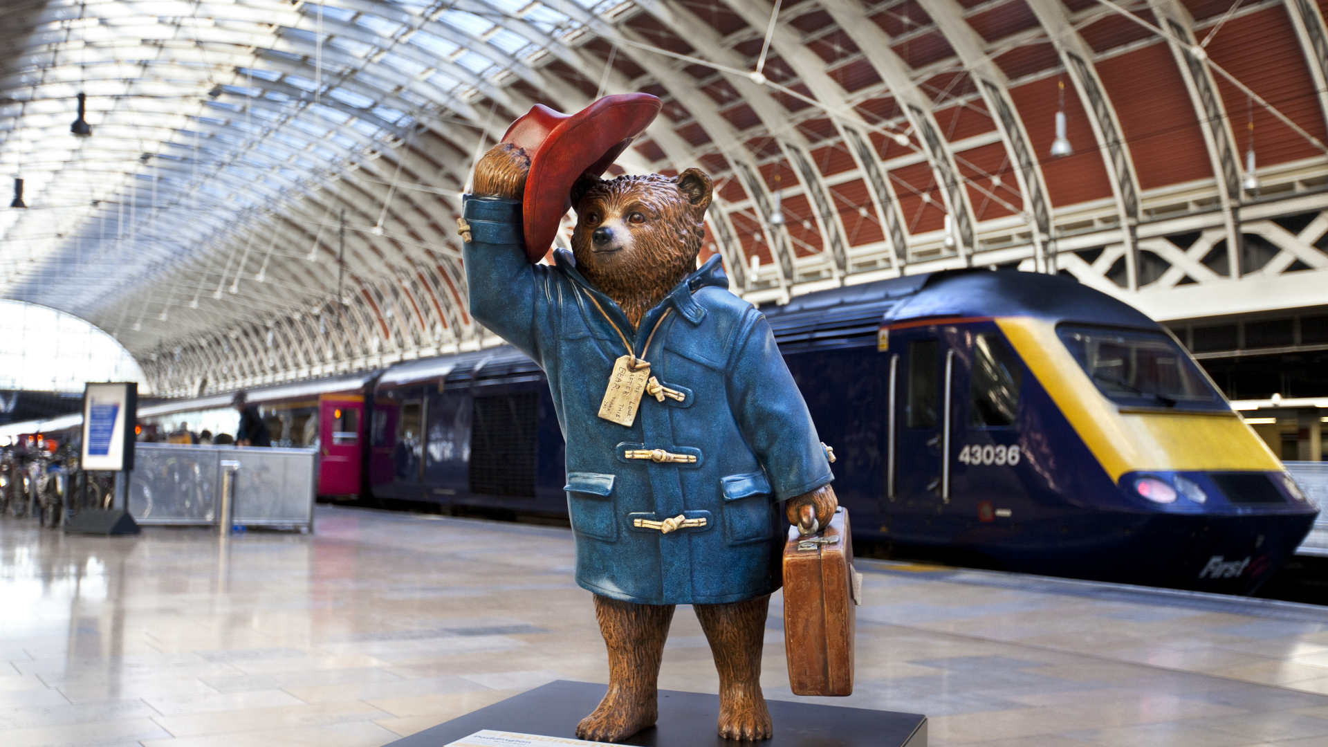 A bronze Paddington Bear statue at Paddington Station. © Chris Dorney/Shutterstock A bronze statue of Paddington Bear in his blue coat holding a briefcase and tipping his red hat on a platform at Paddington station with a train in the background