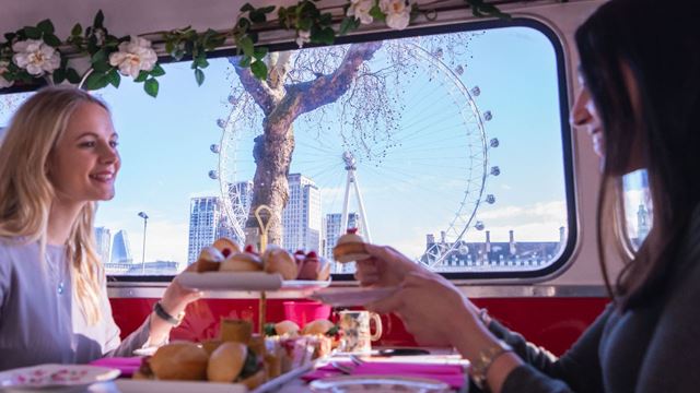 Two women enjoying cakes displayed on a cake stand aboard a bus, with views of the London Eye in the background.