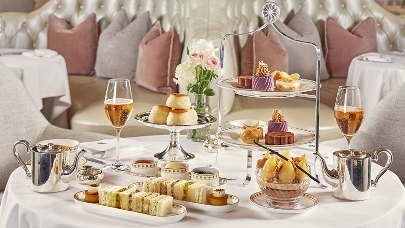Afternoon tea sandwiches, cakes and scones, with glasses of champagne and tea on table with white tablecloth and white seating with pink cushions in the background.