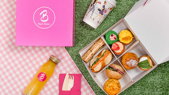 Brigit's Bakery's pink afternoon tea picnic box laid out on grass and pink and white tablecloth, featuring mini sandwiches, cakes and scones, a bottle of orange juice, wooden cutlery and coffee.