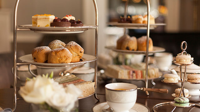 Afternoon tea cakes and scones displayed on two multi-tiered cake stands, with cups of tea and flowers in the foreground.