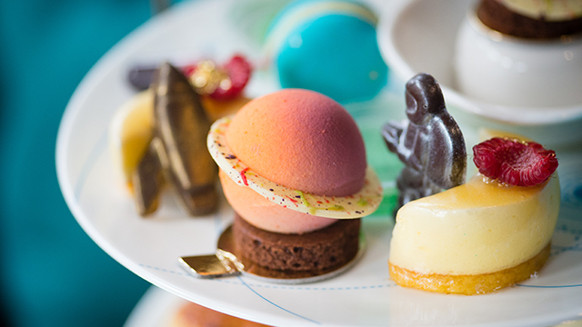 A close-up view of colourful cakes, including one shaped like a planet with rings, sitting on the top tier of a cake stand.