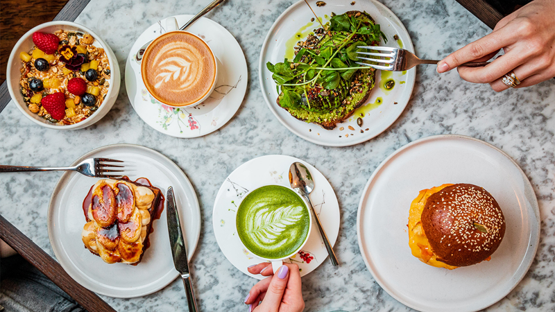 A table spread of brunch dishes including avocado toast, tahini and banana bread, granola, a burger, a latte and matcha