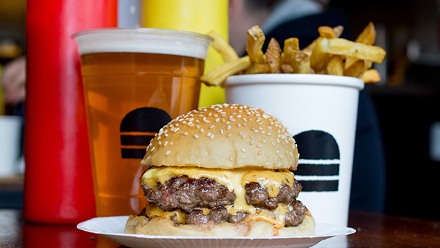 Satisfy your taste buds with a burger, chips and beer © Bleecker. Image courtesy of Bleecker. A burger on a paper plate with a beer, chips and bottles of ketchup and mustard behind