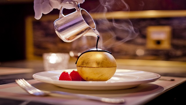 Landscape shot of a gold-covered chocolate dome on a plate, with somebody pouring chocolate sauce from a small jug from above.