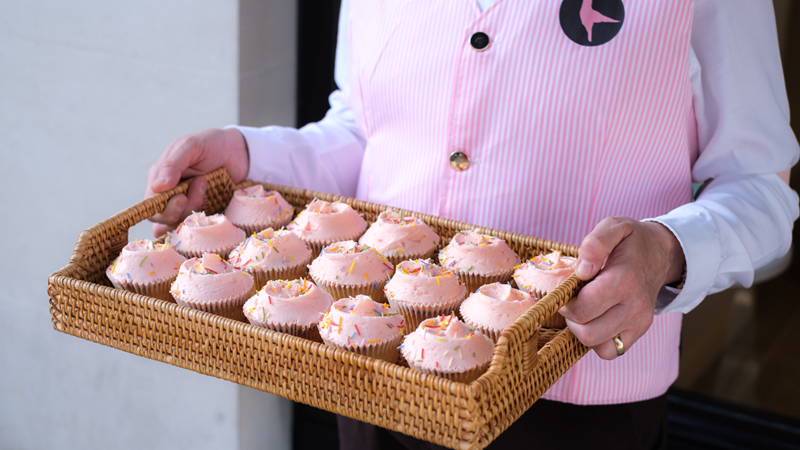 Choose from pretty cupcakes, personalised celebration cakes and seasonal specials at The Hummingbird Bakery. Credit: The Hummingbird Bakery. Image courtesy of The Hummingbird Bakery. A Hummingbird Bakery employee wearing a white shirt and pink waistcoat holds a wooden tray with pink cupcakes topped with sprinkles