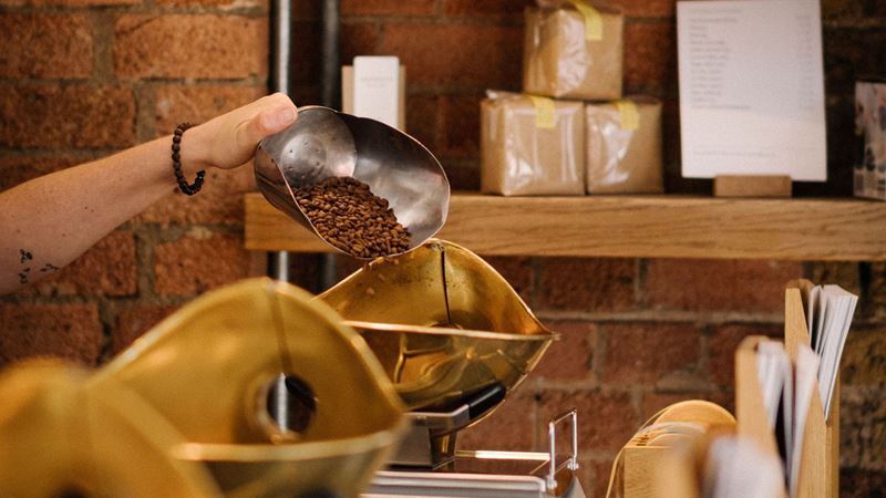 A close up photo of a hand holding a big metal scoop filled with coffee beans being poured into gold weighing scales inside Monmouth Borough