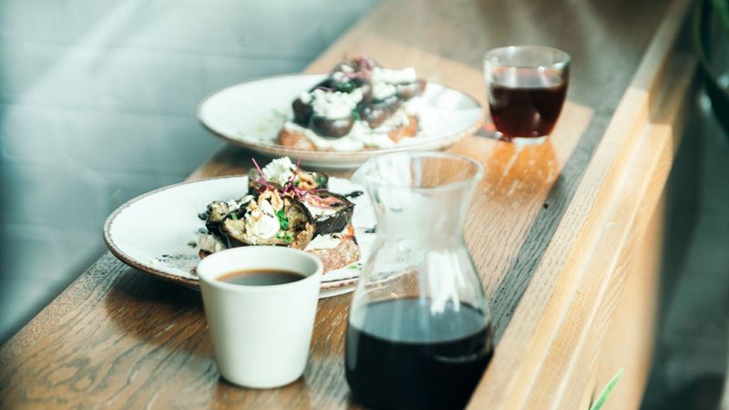 Choose from a menu of tasty dishes to accompany your favourite brew at Prufrock. Credit: Prufrock Coffee. Image courtesy of Prufrock Coffee. A photo of two plates of brunch dishes, a mug of coffee and a glass of coffee with a carafe filled with black coffee on a wooden table inside Prufrock Coffee