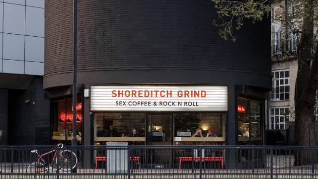 An  espresso bar with a white banner stating shoreditch grind, with red benches outside and a red bike.