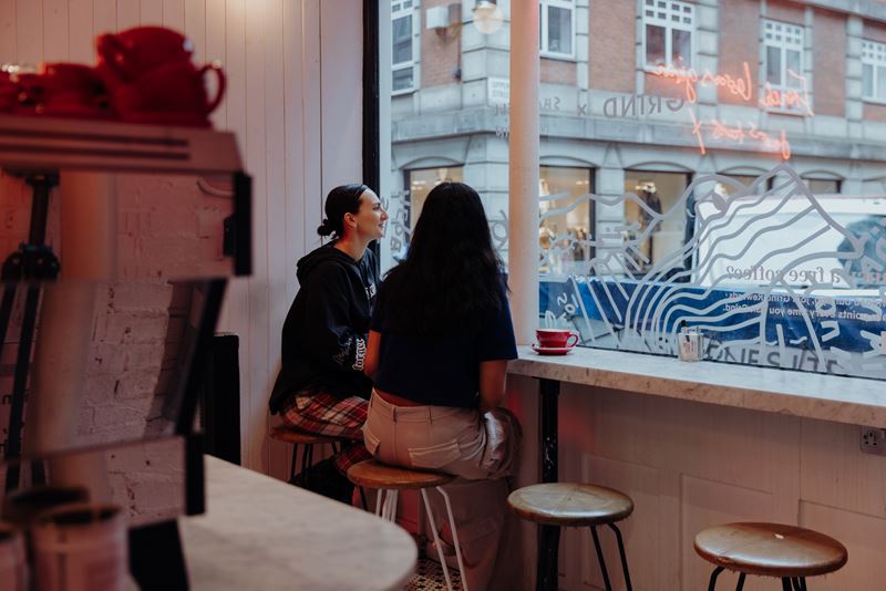 Sip coffee next to the neon lights inside Grind Soho and stay for the evening cocktail menu. Credit: Rebecca Hope. Image courtesy of Soho Grind. A photo of two friends sat on stools inside Soho Grind coffee shop looking out the window onto the street with a coffee machine and neon lights in the background
