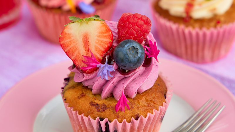 A cupcake on a pink plate with pink icing topped with a blueberry, raspberry half cut strawberry and little edible flowers
