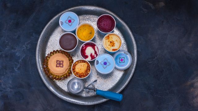 A silver tray that has a variety of ice cream tubs filled with different flavours and a large blue ice cream scoop.