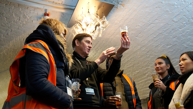 Learn about the beermaking process during a Fuller's Brewery Tour. Credit: Fuller's Brewery. Image courtesy of Fuller's Brewery. A tour guide holds up a glass of freshly brewed beer while talking to a group wearing orange hi-vis jackets on a Fullers Brewery tour
