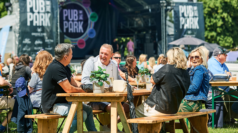 Enjoy an unforgettable day out in the sun at Pub in the Park with plenty of delicious food and drink. Credit: G Bush. Image courtesy of Pub in the Park. A group of people sat on a bench outside at Pub in the Park