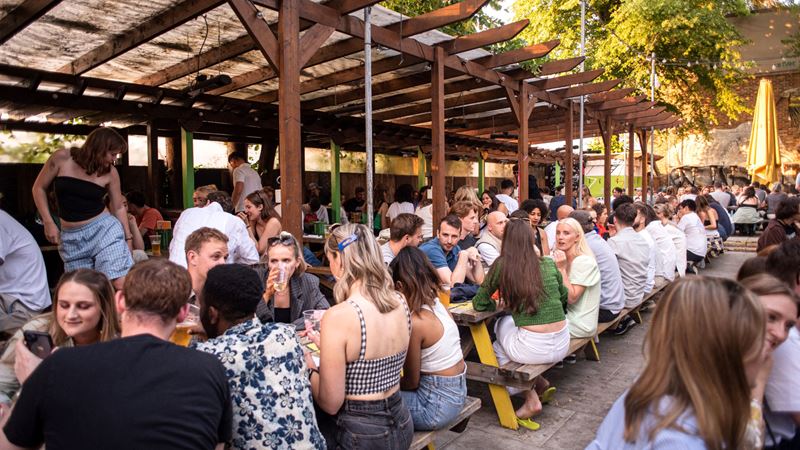 Soak up the sunshine from the Duke of Edinburgh's huge garden. Credit: Nina Pennick. Image courtesy of Rose Pubs. A photo of crowds of people sat on picnic benches in the Duke of Edinburgh's outdoor beer garden with wooden canopies and greenery in the background as the sun shines down
