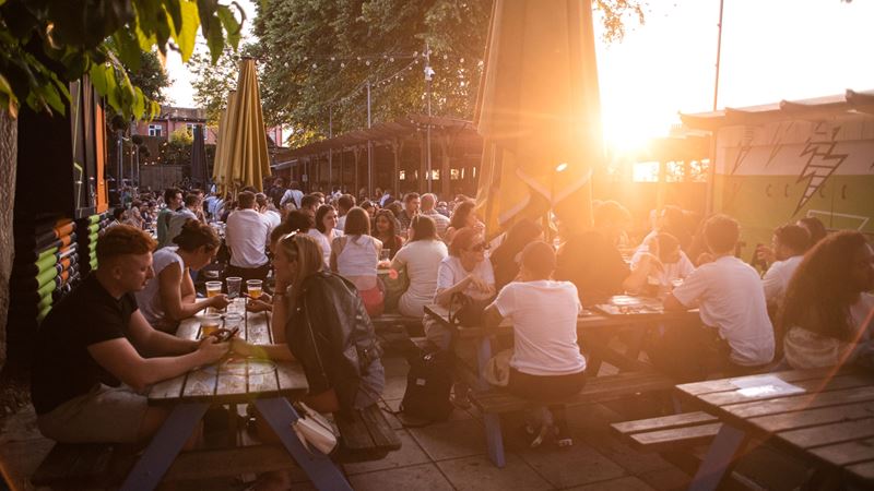 A photo of the sun low in the sky shining into the Duke of Edinburgh pub garden as lots of people sit on picnic benches chatting, smiling and sipping drinks