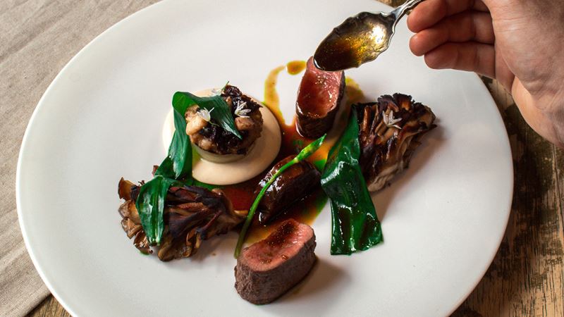A photo of a muntjac with ale dish at The Harwood Arms featuring medium rare slices of muntjac, wilted wild garlic leaves and a man's hand dripping an ale sauce over the dish with a spoon