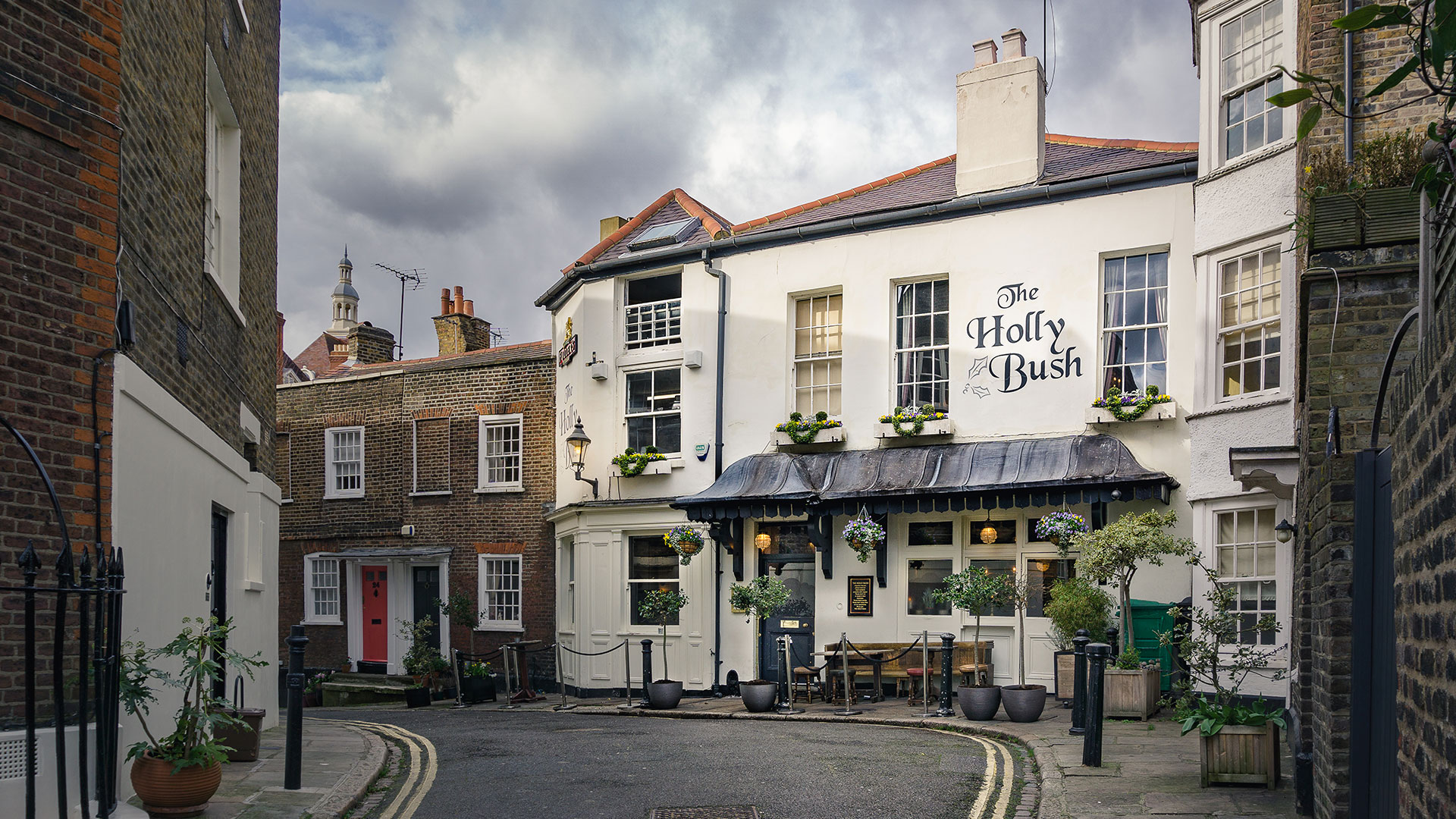 Visit cosy pub The Holly Bush in quaint Hampstead. Credit: The Holly Bush. Image courtesy of The Holly Bush. The facade of The Holly Bush pub in Hampstead and the street leading up to it on a clear day.
