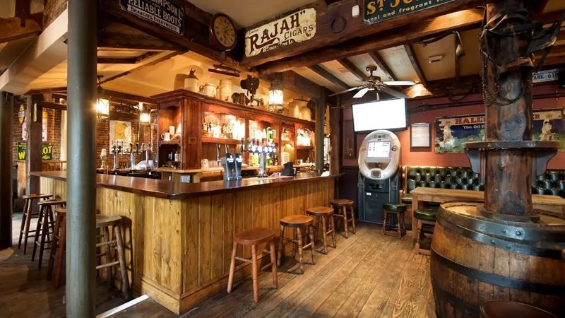 Watch live sports or play darts with pizza and a pint at The Alexandra in Clapham. Credit: The Alexandra. Image courtesy of Greene King. The wooden bar area at The Alexandra pub in Clapham, with stools, drinks and a TV screen in the background.