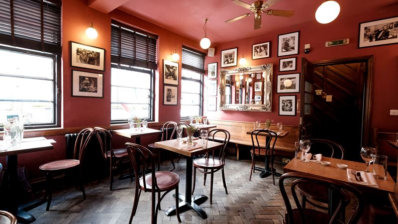 Step inside the bohemian French House pub in Soho. Credit: Peter Clark. Image courtesy of The French House. Warm-coloured interior of The French House pub in Soho, with small, dark wooden tables and chairs, pictures on the walls, wooden parquet floors and dim lighting.