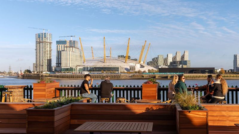Pull up a chair and enjoy a pint at The Gun's riverside garden. Credit: The Gun. Image courtesy of The Gun. A photo of groups of people sat on stools in the riverside garden at The Gun with views of the river Thames and the south bank with The O2 in the distance