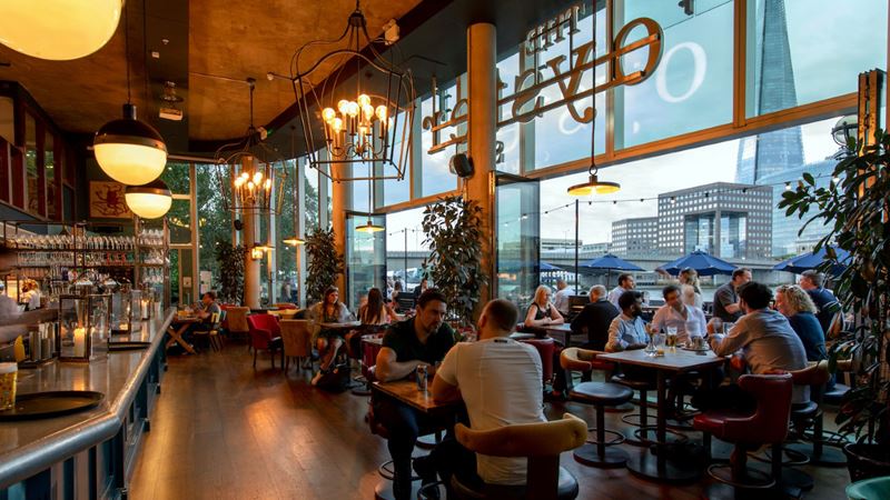 Admire the riverside views from the floor-to-ceiling windows at The Oyster Shed. Credit: Oyster Shed at Young's Pubs. Image courtesy of The Oyster Shed, A photo of The Oyster Shed interior with people sat next to floor-to-ceiling windows with views of the south bank of the Thames including The Shard