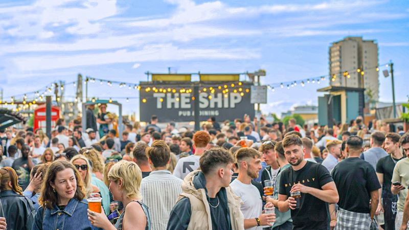 Join the crowds that flock to The Ship in Wandsworth for al fresco riverside drinks. Credit: Ella Umney. Image courtesy of The Ship. A photo of crowds of people chatting, smiling and with drinks in hand in the garden at The Ship with fairy lights and a blue sky above