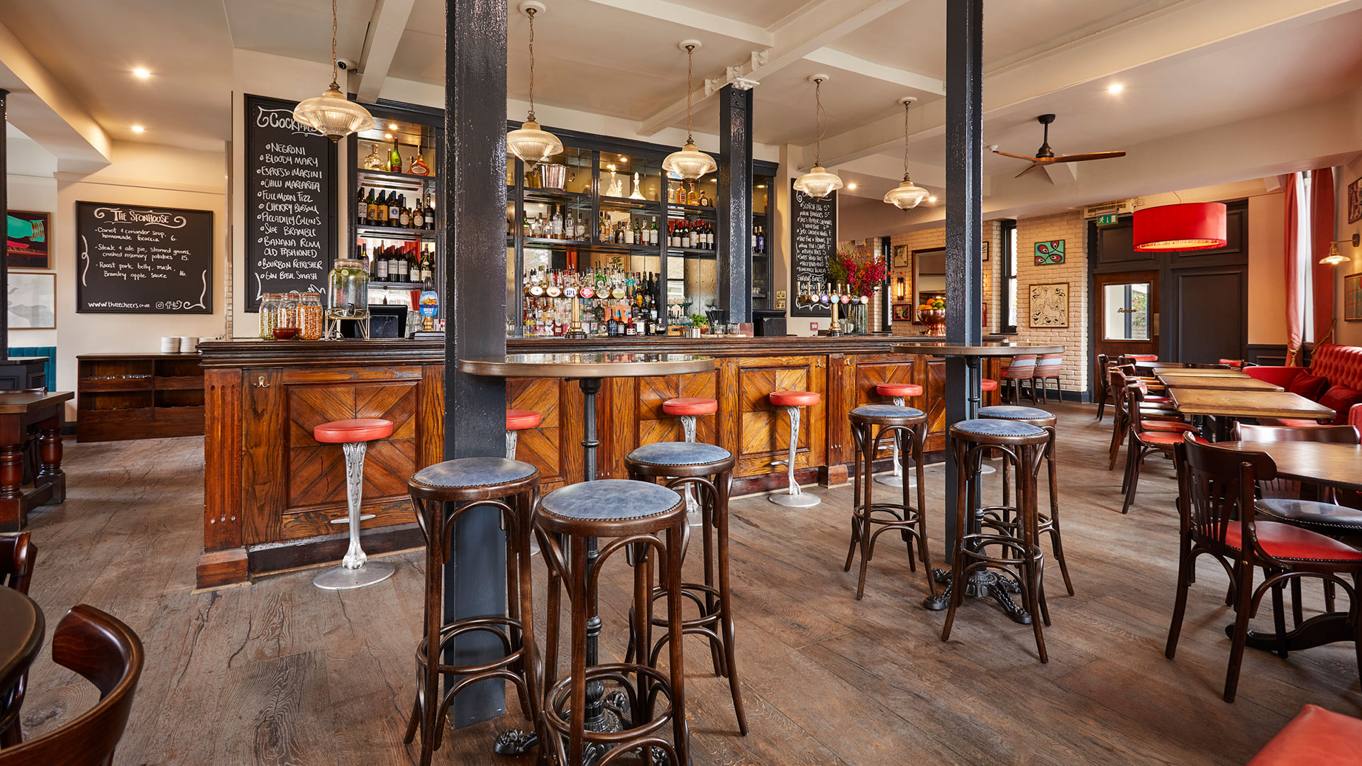 Settle in and enjoy a hearty meal and a pint at this local favourite The Stonhouse. Credit: The Stonhouse. Image courtesy of Three Cheers Pub Co. Inside The Stonhouse pub in Clapham, featuring wooden floors and tables, red and blue seats, a wooden bar area with drinks lined on shelves in the background.