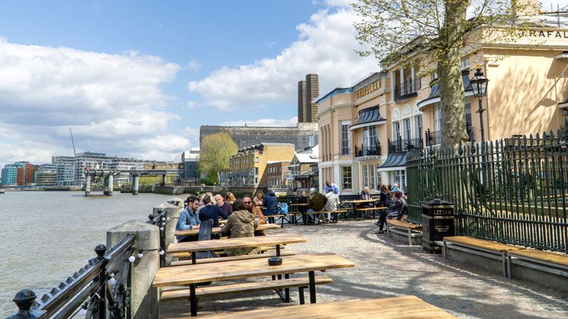 Take in the city skyline from the water's edge seating at the Trafalgar Tavern. Credit: Shutterstock. Image courtesy of Shutterstock. A photo of the exterior of the Trafalgar Tavern with people sat on the riverside benches with the Thames in front of the pub